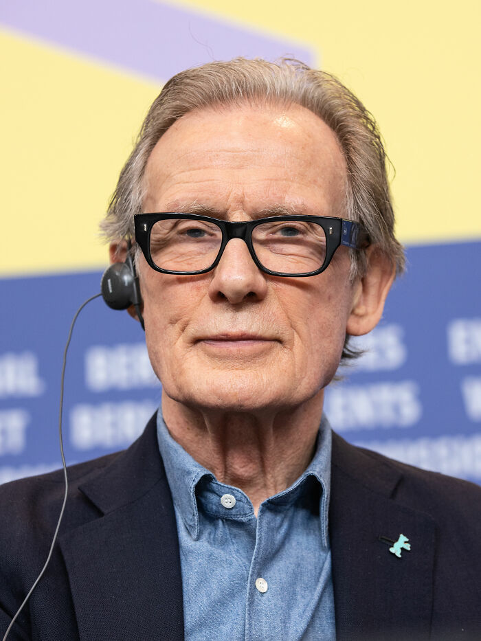 Bill Nighy wearing glasses and a headset in a formal setting, related to his bio and career highlights.