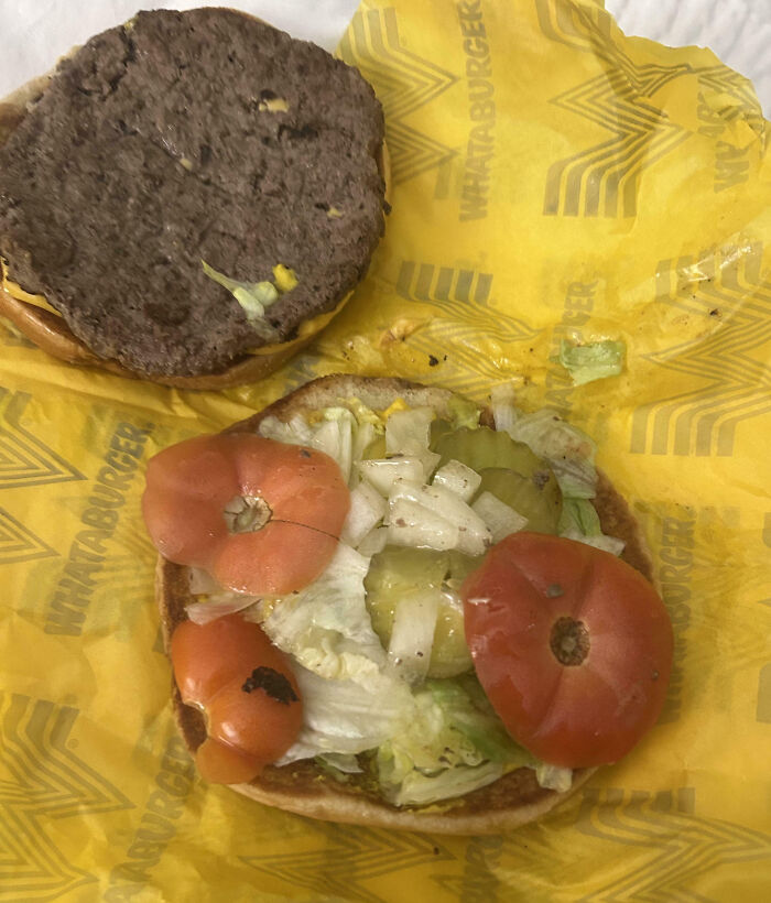 Burger with uneven tomato slices, lettuce, pickles, and a patty on yellow fast food wrapper showing outrageous meal difference.