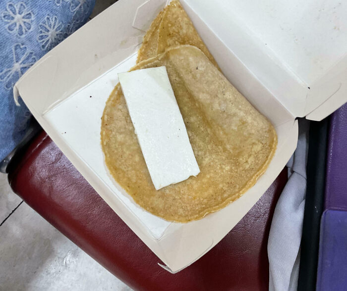 A single thin taco shell with a plain rectangular piece of white cheese inside in a takeout box, showing outrageous meal disparity.