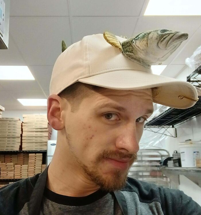 Man in a food service job wearing a white cap with a stuffed fish attached, posing in a kitchen storage area.
