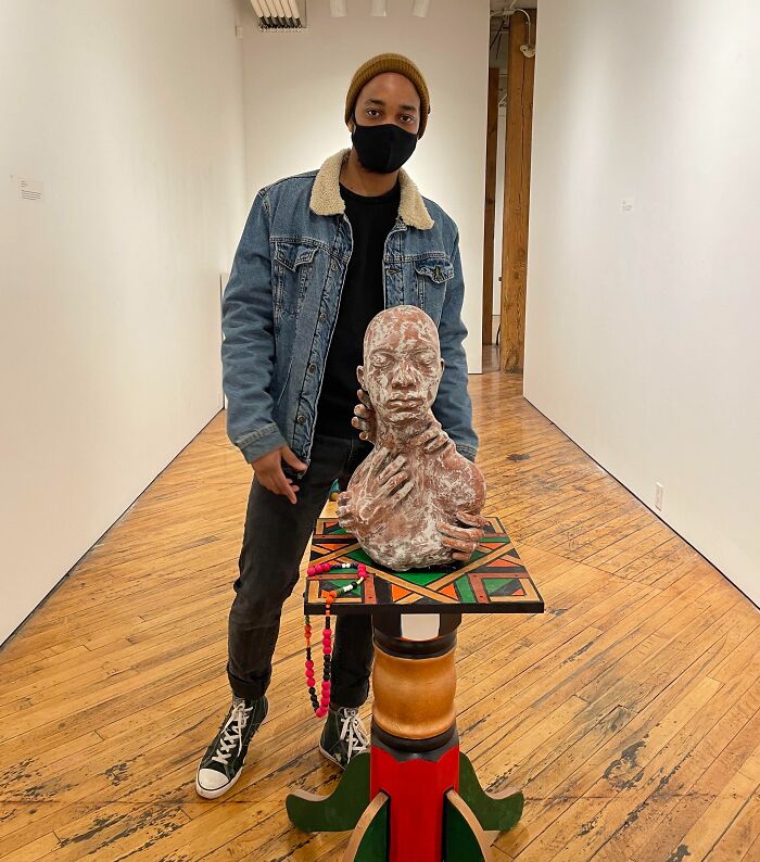 Artist in a gallery wearing a mask standing next to a sculpted bust on a colorful pedestal showcasing unique job photos.