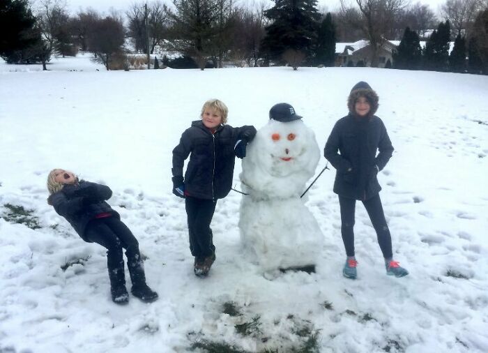 Three kids posing with a snowman in a snowy yard, capturing a hilariously awkward family Christmas photo moment.