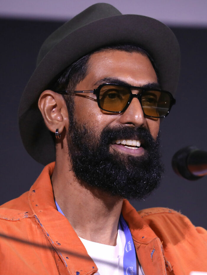 Rana Daggubati wearing sunglasses and a hat, speaking at an event while smiling and dressed in an orange jacket.