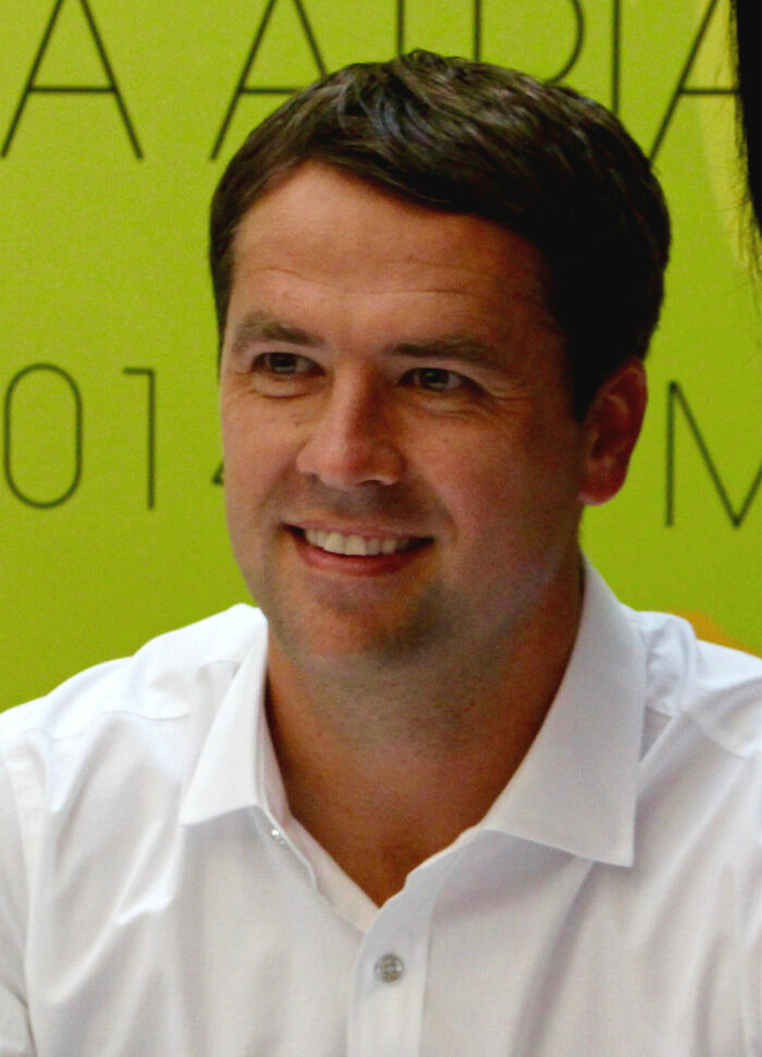 Michael Owen smiling, wearing a white shirt, in a casual setting focused on bio and career highlights.