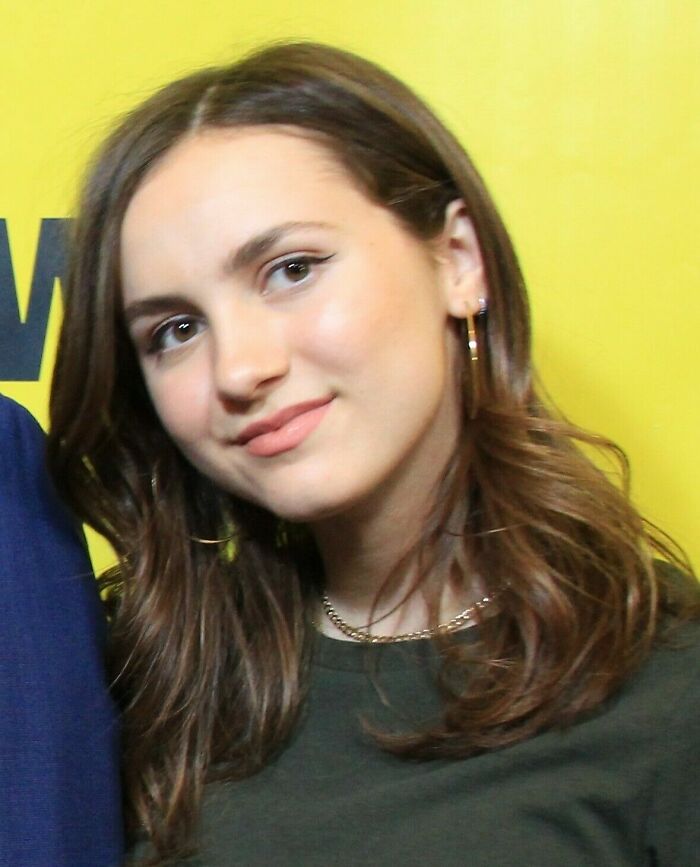 Maude Apatow smiling with wavy brown hair and hoop earrings against a yellow background at an event.
