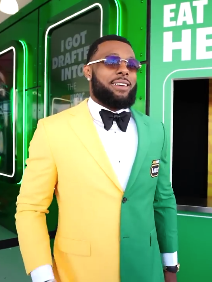 Man wearing a half yellow, half green suit with bow tie, sunglasses, smiling at a green promotional event setup.