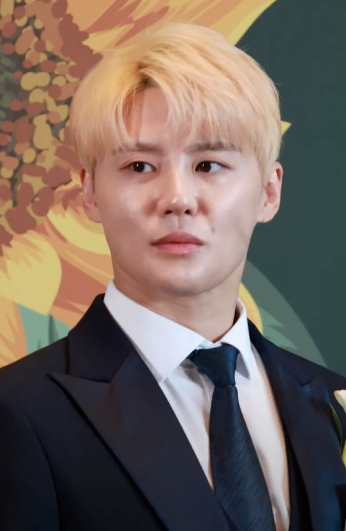 Kim Junsu in a dark suit and tie, with blonde hair, standing in front of a colorful sunflower background.