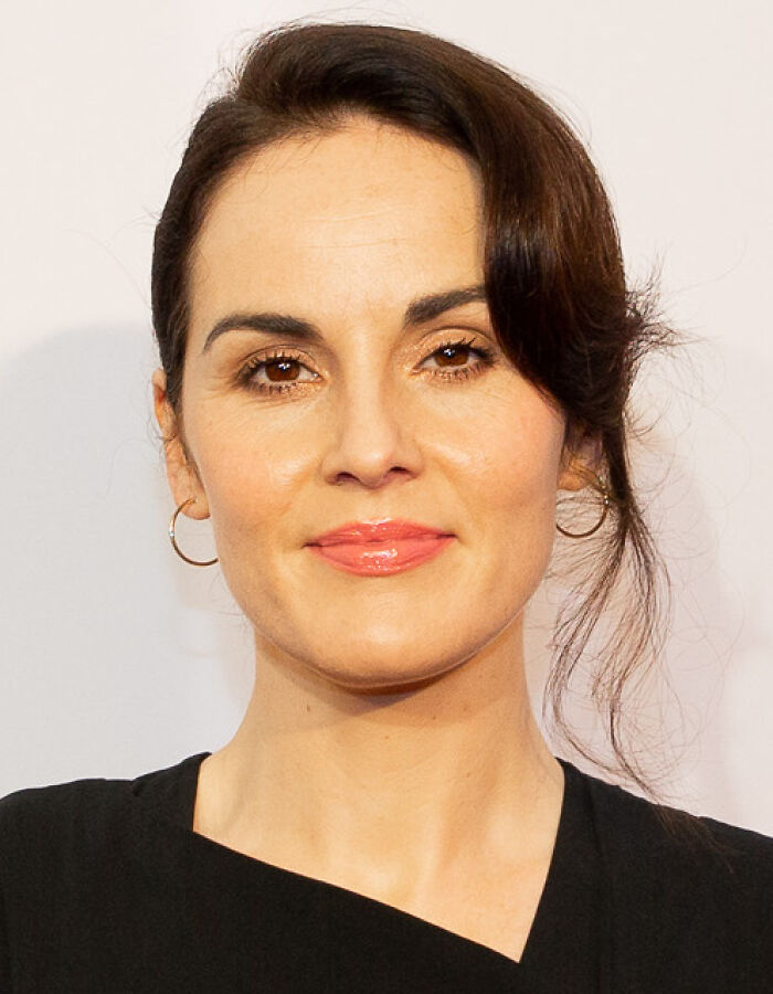 Michelle Dockery smiling at an event with hoop earrings and natural makeup, showcasing her bio and career highlights.
