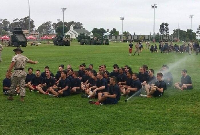 Group of young men sitting on grass during military training with a sprinkler spraying water, capturing a funny perfect time moment.