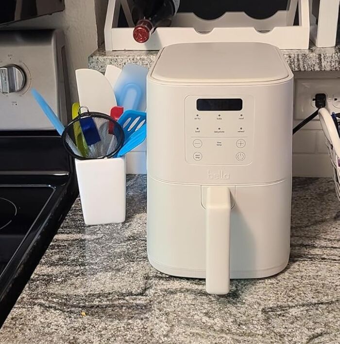 White Bella air fryer on kitchen counter next to blue kitchen utensils, a stove, and a wine bottle rack in the background.