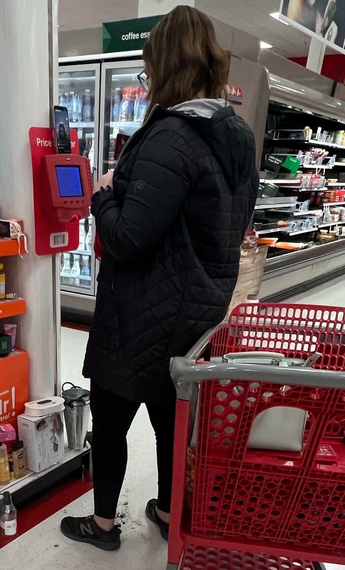 Woman in a black jacket scanning items in a Walmart store aisle with a red shopping cart nearby and refrigerated shelves behind.