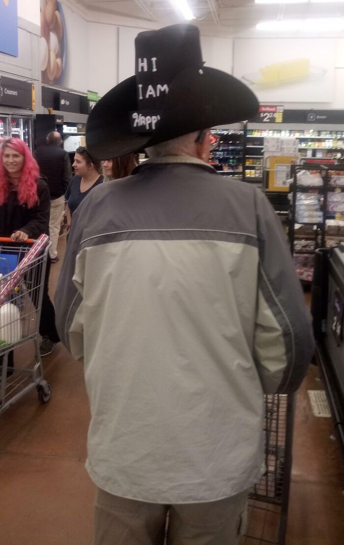 Man wearing oversized cowboy hat with message inside Walmart aisle among shoppers and carts.