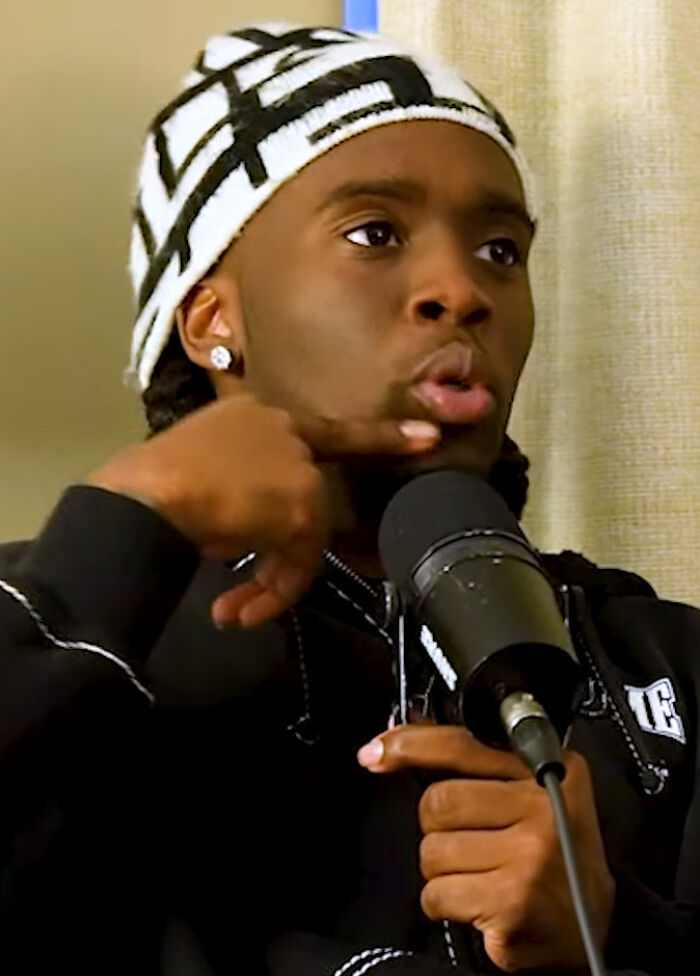 Kai Cenat wearing a black hoodie and patterned beanie speaking into a microphone during an indoor interview session.