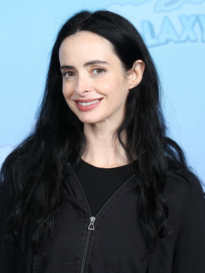 Krysten Ritter with long black hair, wearing a black zip-up jacket, smiling against a light blue background.