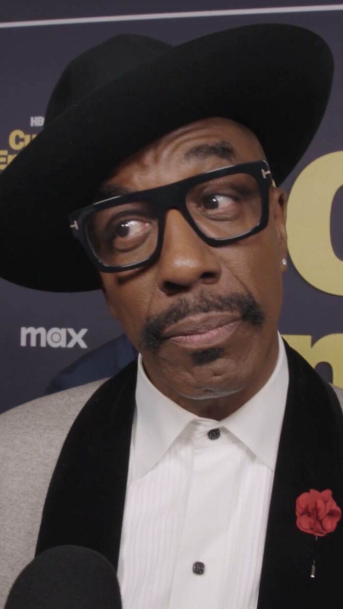 J. B. Smoove wearing black hat and glasses, dressed formally with a red flower lapel pin at an event.