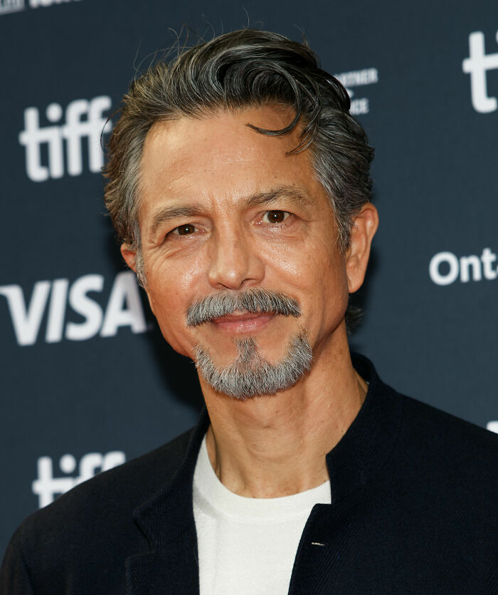 Benjamin Bratt smiling at a film event, wearing a black jacket and white shirt against a dark background.