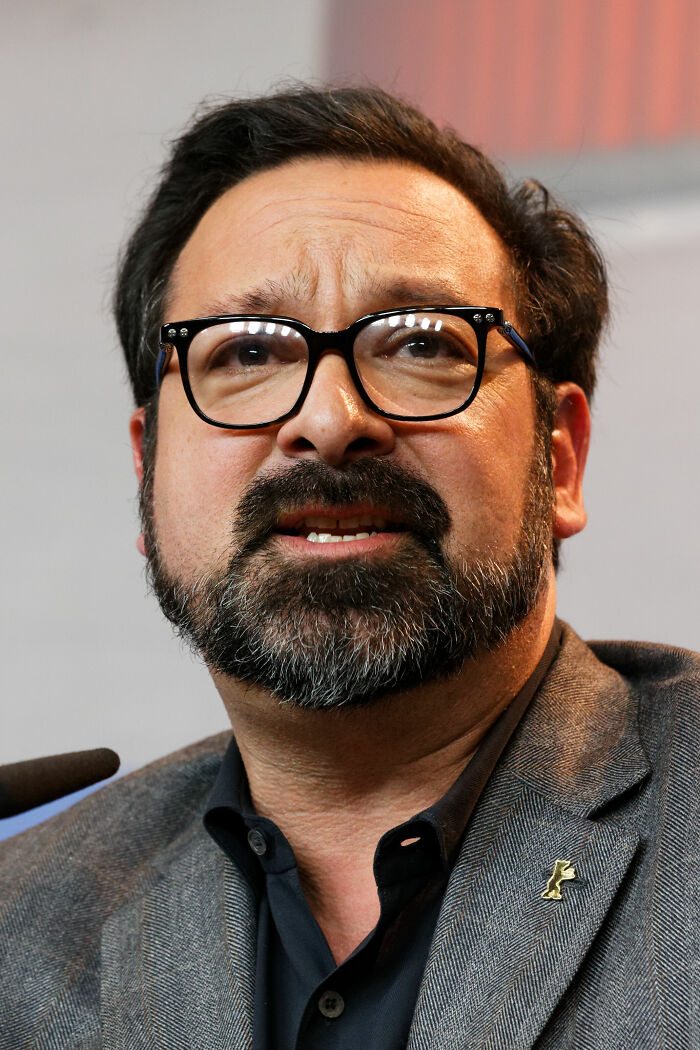 James Mangold wearing glasses and a grey jacket speaking at an event, highlighting his bio and career achievements.