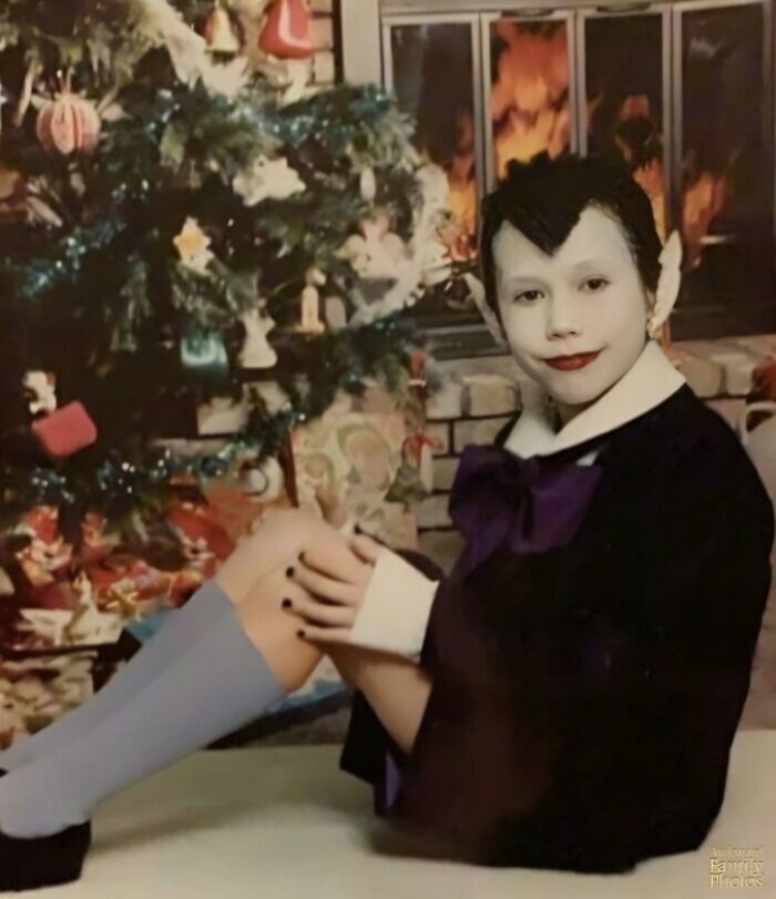 Child in Halloween vampire costume poses awkwardly by Christmas tree in hilariously awkward family Christmas photo.