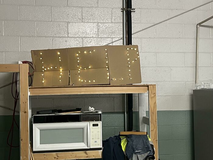 Cardboard sign with lights spelling help on a wooden shelf above a microwave, creative workplace Christmas decoration.