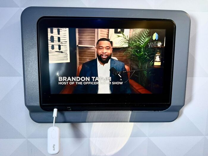 Tablet screen showing a podcast host speaking indoors, with a small white AirFly device plugged in below the display.