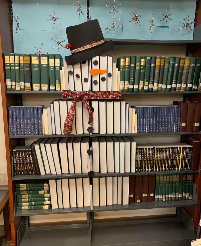 Creative workplace Christmas decoration showing a snowman made from arranged books with hat and scarf on a bookshelf.