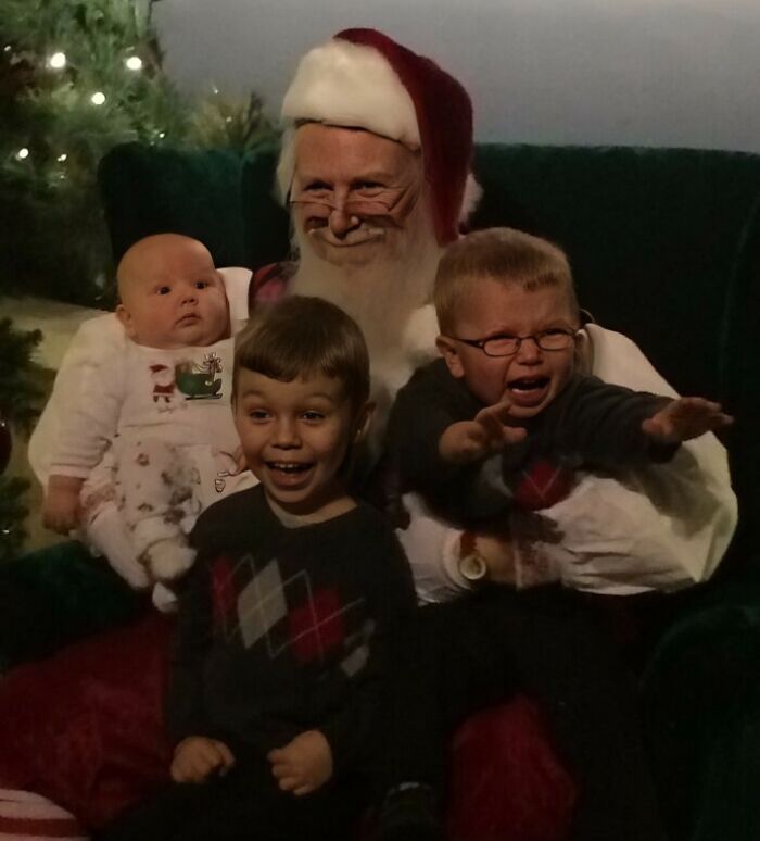 Crying child and smiling kids sitting with Santa Claus during awkward family Christmas photo moment.