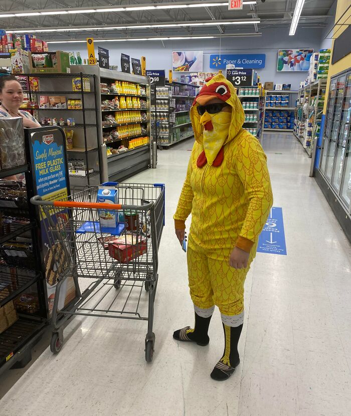Person dressed in a yellow chicken costume shopping at Walmart aisle with a cart and another shopper nearby.