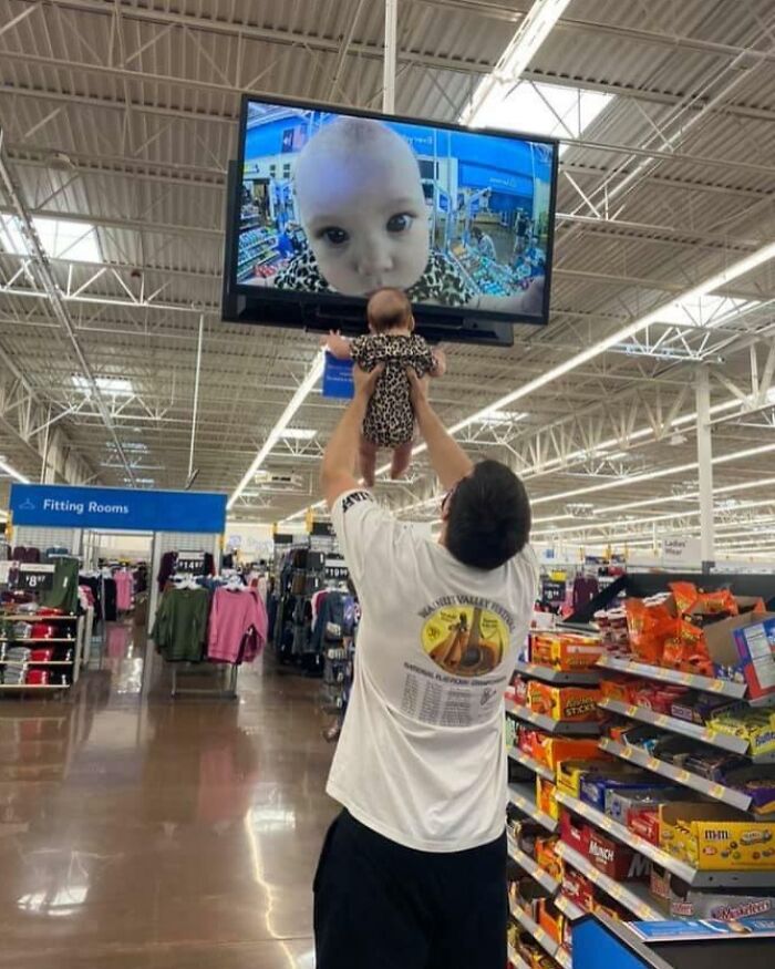 Man holding baby in Walmart aisle as baby’s face is shown magnified on security monitor in the store.