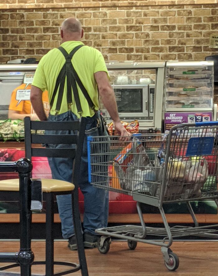 Man in bright yellow shirt with suspenders pushing a shopping cart filled with groceries inside a Walmart store.