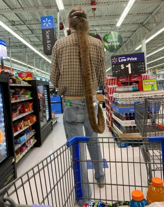 Man with extremely long fuzzy ponytail shopping inside Walmart aisle near candy and water displays.