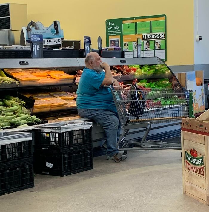 Man sitting inside Walmart produce section with shopping cart filled with groceries, surrounded by vegetables and packaged meats.