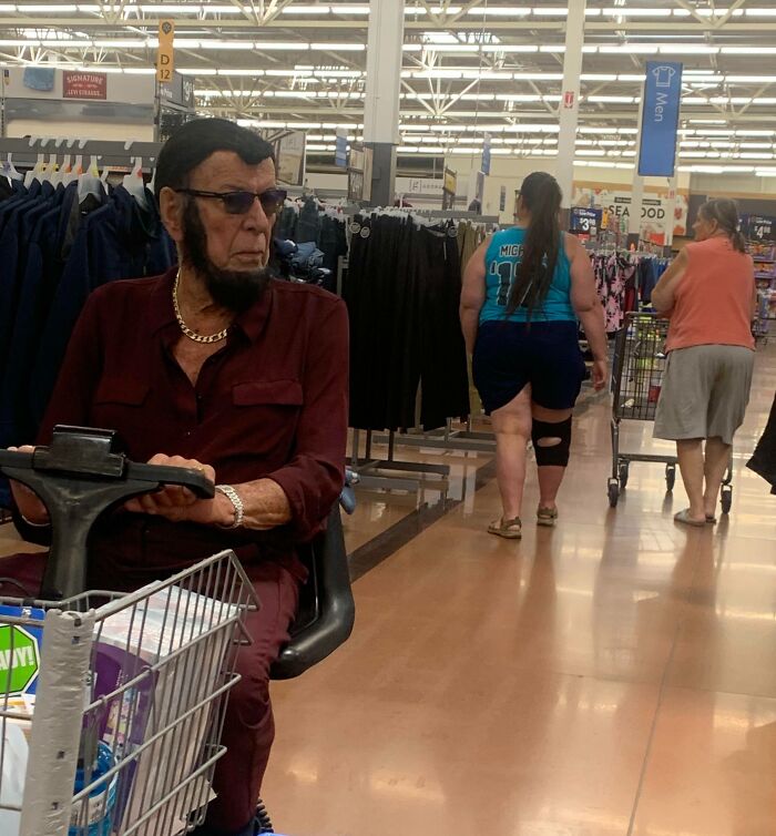 Man with unique hairstyle and beard riding mobility scooter inside Walmart store aisles surrounded by shoppers and clothing racks.