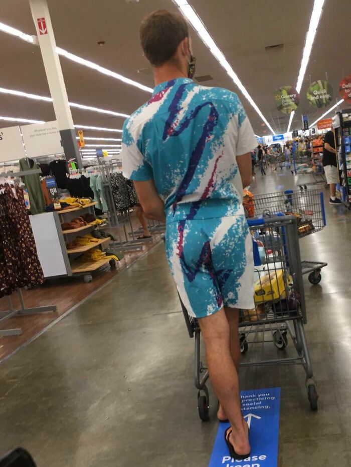 Man in a colorful outfit pushing a shopping cart inside Walmart aisle filled with clothing and grocery items.