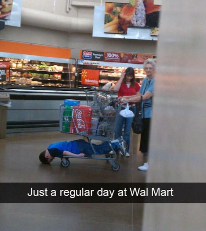 Child lying flat inside a Walmart shopping cart with other shoppers nearby in a grocery aisle.