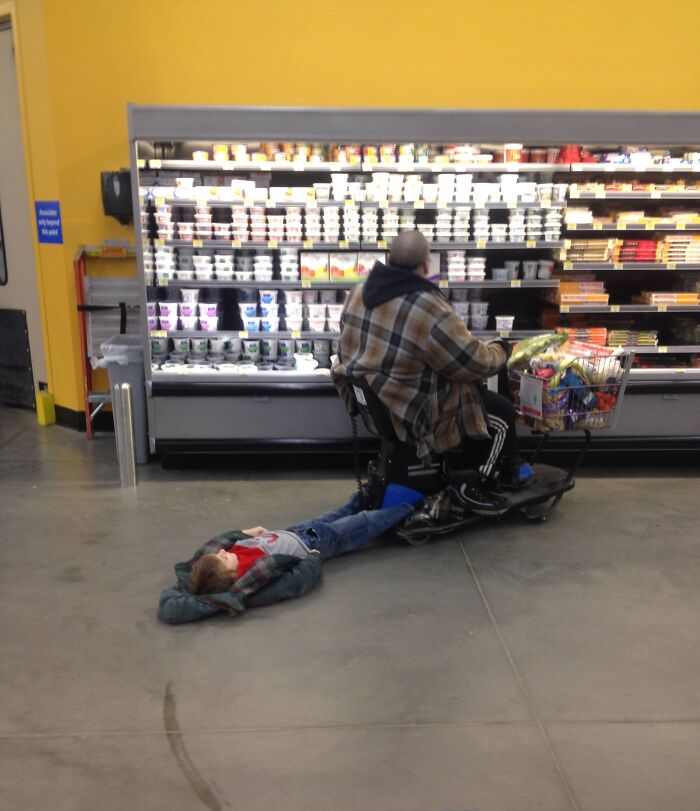 Man on mobility scooter at Walmart with child lying on floor holding onto scooter footrest near dairy aisle