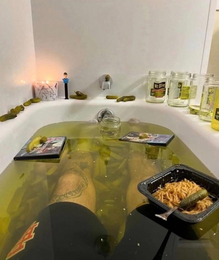 Person relaxing in a bathtub filled with pickle juice and surrounded by pickles, noodles, and empty jars in a humorous food fail.