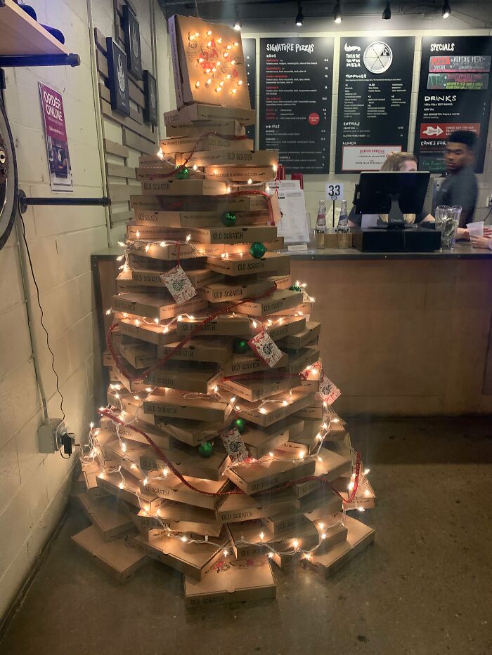Creative workplace Christmas decoration made from stacked pizza boxes wrapped in lights and ornaments at a pizza shop counter.