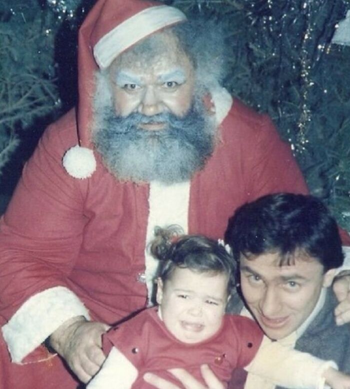 Awkward family Christmas photo with a distressed child, a man smiling, and a Santa Claus with a gray beard.