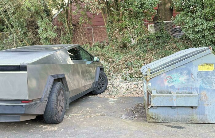 Matte black futuristic truck parked near a graffiti-covered dumpster in a wooded outdoor area, capturing funny pictures weirdness.