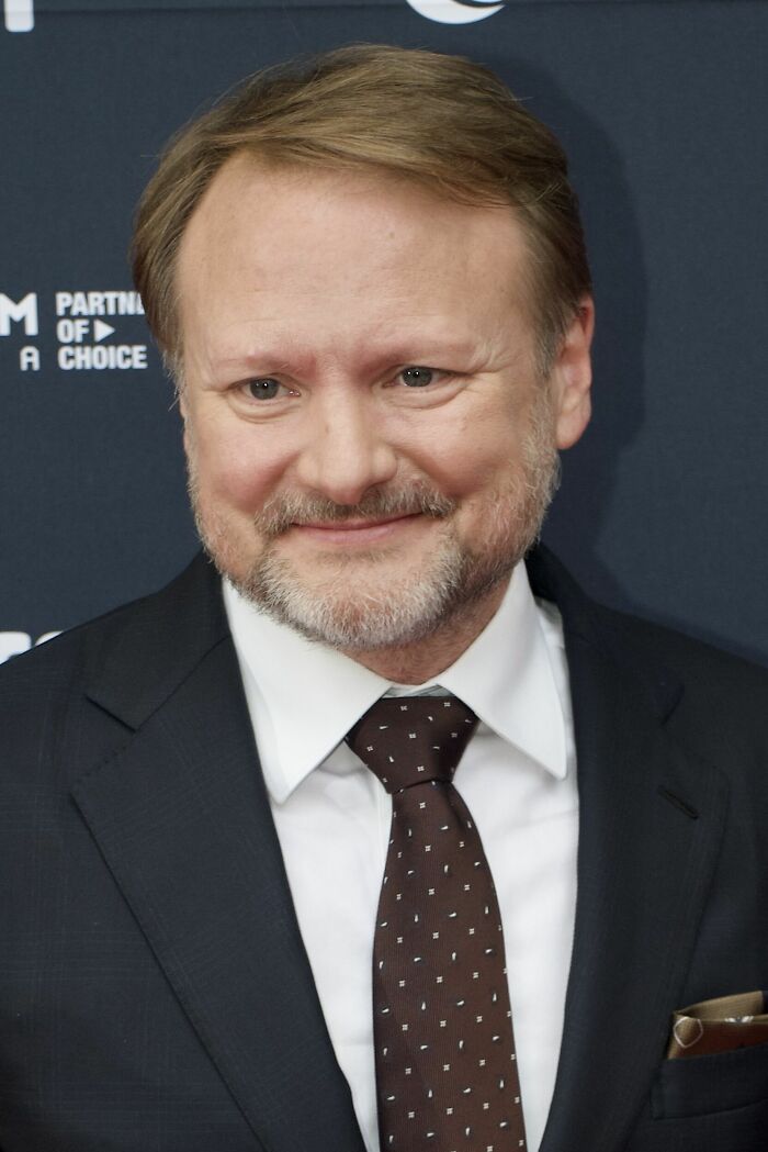Rian Johnson smiling at an event, wearing a dark suit and patterned tie, highlighting his bio and career achievements.