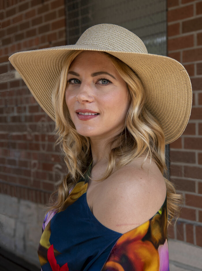 Katheryn Winnick wearing a wide-brimmed hat and colorful off-shoulder top, posing outdoors by a brick wall.