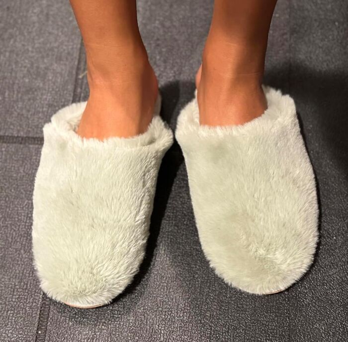 Feet wearing soft, fluffy slippers indoors on a dark textured floor, ideal gifts for the person who has everything.