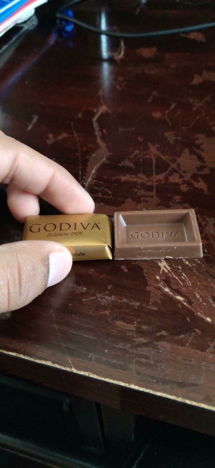Small Godiva chocolate square next to its gold wrapper on a scratched dark wooden surface showing expectations vs reality.