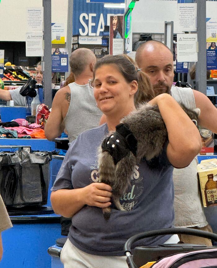 Woman holding a raccoon inside a Walmart store with other shoppers and checkout counters in the background.
