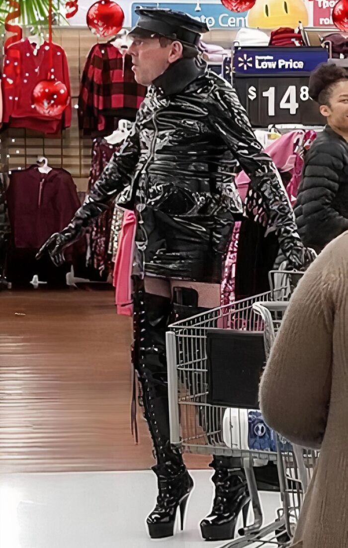Person in shiny black leather outfit and high heels pushing a cart inside Walmart, dressed unusually for the store environment.