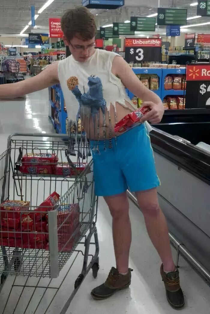 Young man wearing a torn Cookie Monster shirt shops at Walmart, holding snacks with a cart full of similar items in an aisle.