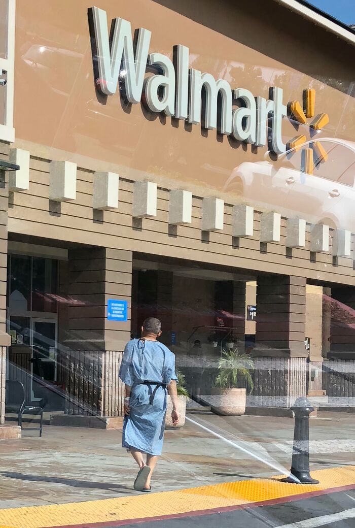 Person in hospital gown walking outside Walmart entrance with sprinklers spraying water on the sidewalk in front of the store