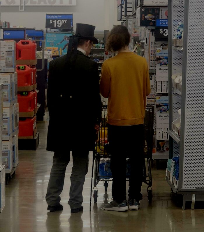 Two shoppers in a Walmart aisle, one wearing a top hat, pushing a cart amid store products and displays.