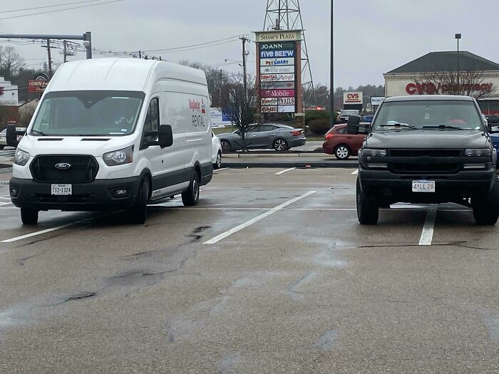 Two vehicles parked across multiple spaces in a wet shopping center lot, illustrating next-level jerks.