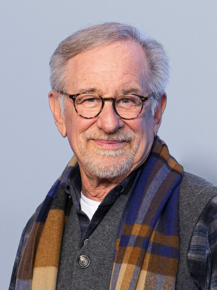 Steven Spielberg wearing glasses and a scarf, smiling in a close-up portrait with a light blue background.